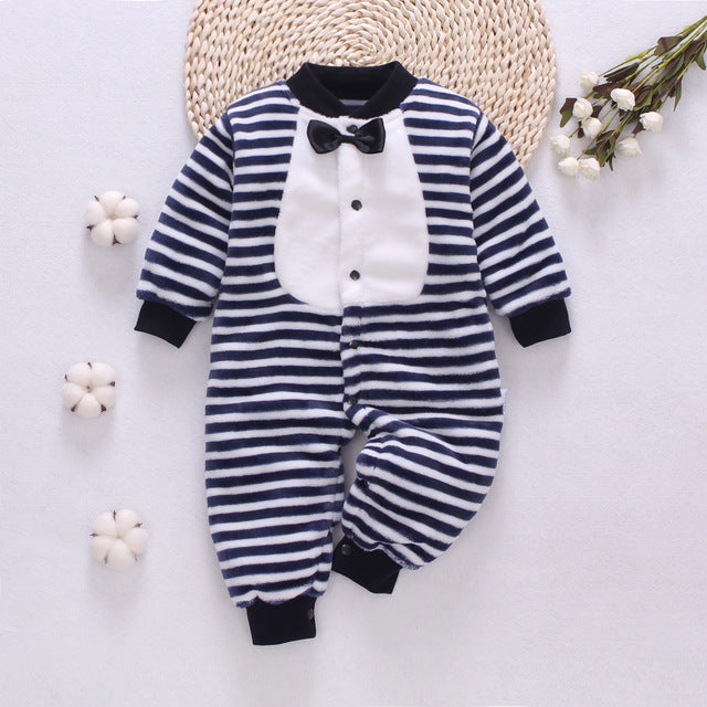 Striped navy and white baby romper with white tuxedo design and black bow tie. Set on a woven mat with cotton flowers and white blossoms nearby.