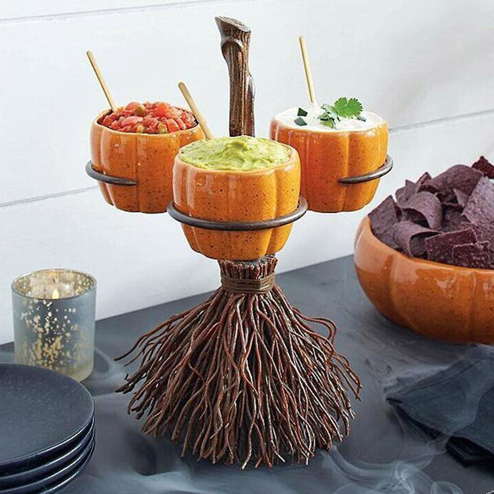 A whimsical broomstick holder with three pumpkin-shaped bowls containing salsa, guacamole, and sour cream. Nearby, a bowl holds blue corn tortilla chips, and a candle is in the background.