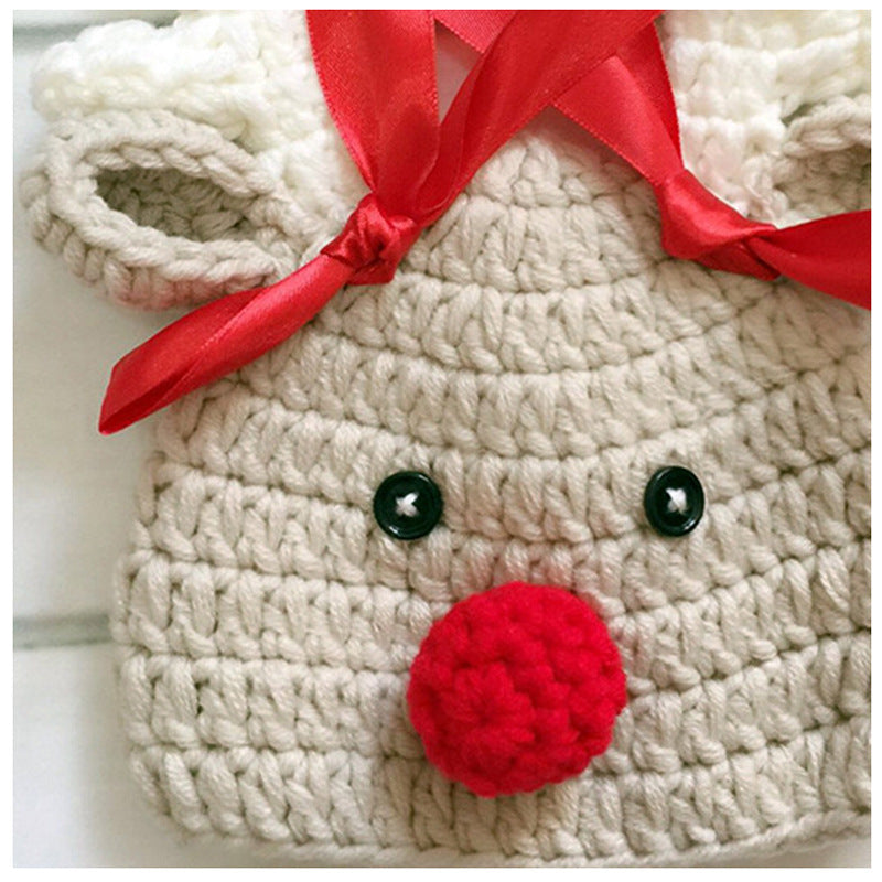 Crocheted reindeer hat with black button eyes, a red nose, and red satin ribbon. White and beige yarn forms the antlers and ears, creating a festive design.