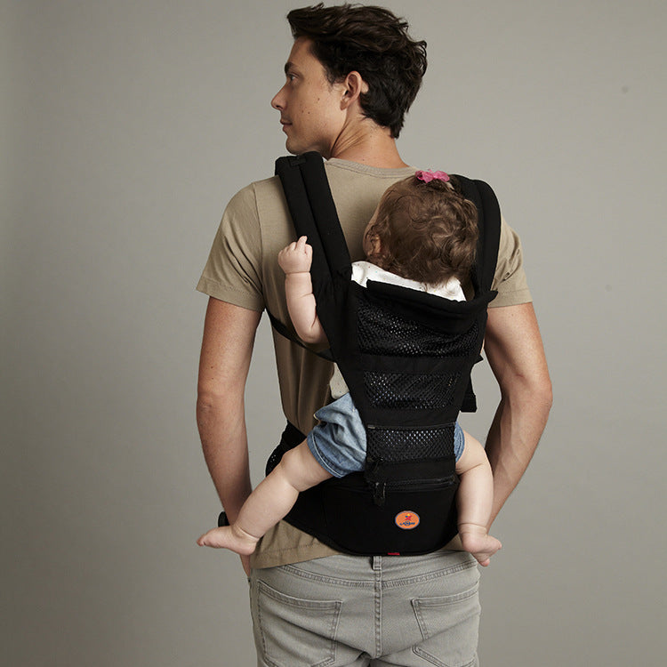 Man wearing a beige t-shirt carries a baby in a black mesh baby carrier on his back. The baby, wearing denim shorts, has a hair bow and rests comfortably against him.