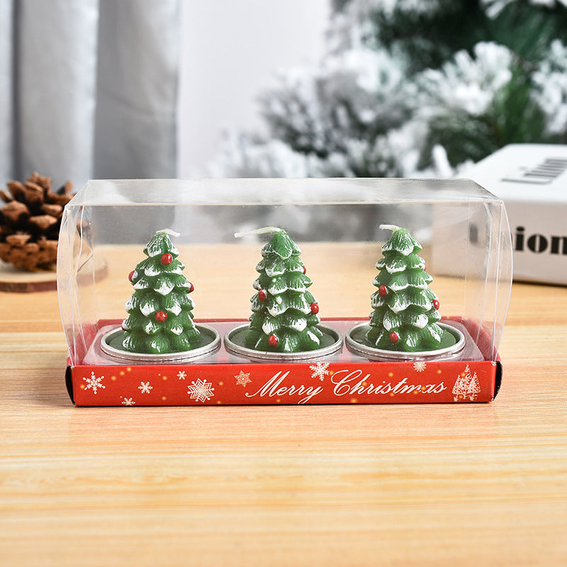 Three decorative Christmas tree candles in a box with a red "Merry Christmas" label. The candles feature green trees with white tips and red ornaments, sitting on a wooden table.