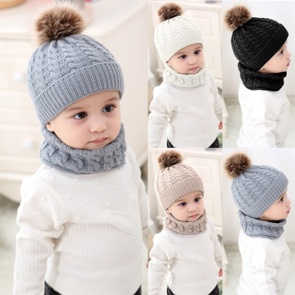 A toddler wearing knitted hats and matching scarves in different colors: gray, cream, black, and beige, each with a fluffy pom-pom, standing in a light room.