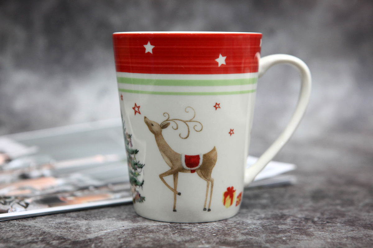 Festive mug with a red border, adorned with stars, featuring a reindeer wearing a red saddle, a Christmas tree, and wrapped gifts, set against a blurred grey background.