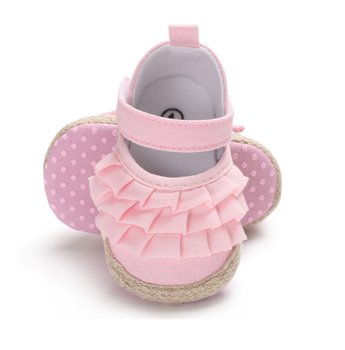 Pink baby shoes with ruffled fabric and a strap, featuring polka dot soles and woven espadrille-style edges, displayed with one shoe upright and the other laying flat.
