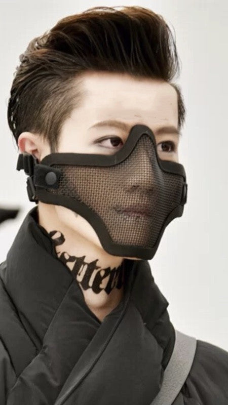 A person with styled hair wears a black mesh face mask and a dark jacket. There is a bold, tattoo-like design on the side of the neck. The background is neutral and blurred.