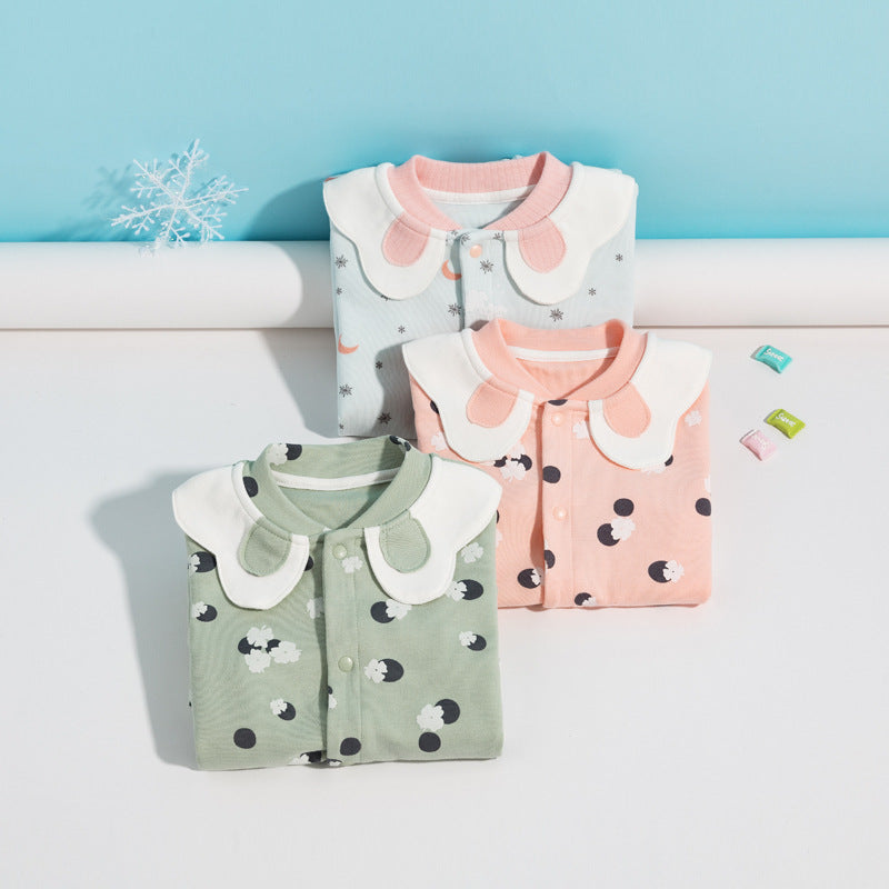 Three folded baby onesies with Peter Pan collars in green, pink, and blue with playful patterns. A snowflake decor and small candies are arranged nearby on a light surface.