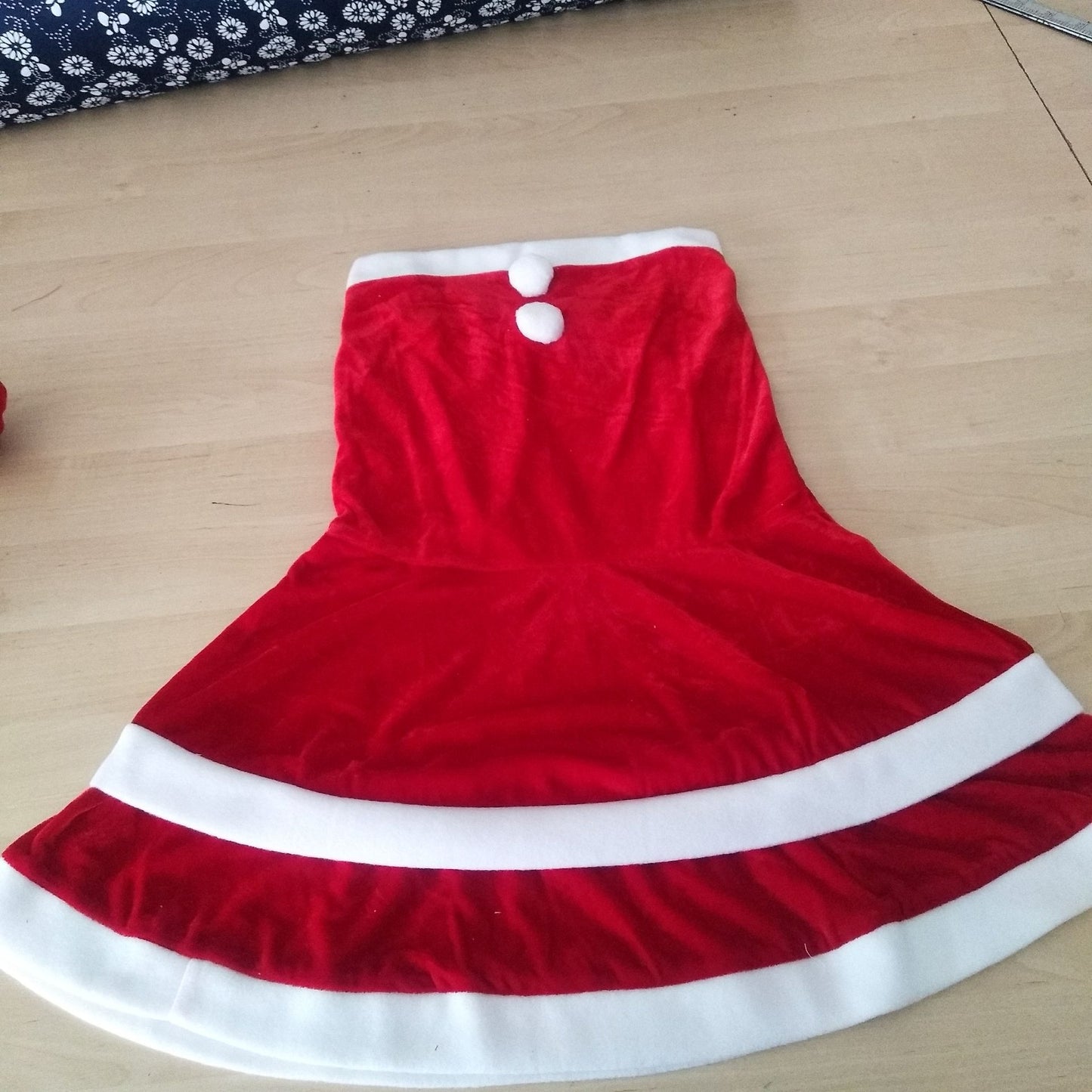Red velvet Christmas tree skirt with two white pom-poms and white borders, laid out on a wooden floor.