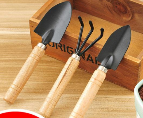Three gardening tools with wooden handles, including two trowels and a hand rake, are placed in front of a wooden box on a wooden surface.