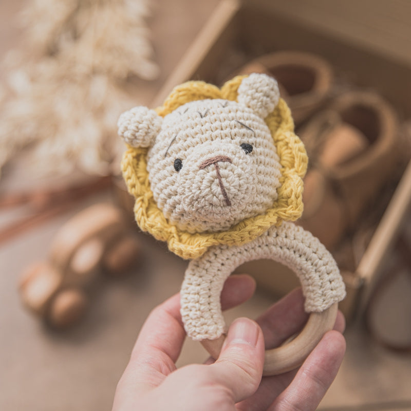 Hand holding a crocheted lion rattle toy with a wooden ring and a yellow mane. Background includes a blurred wooden toy car and baby shoes in a box.