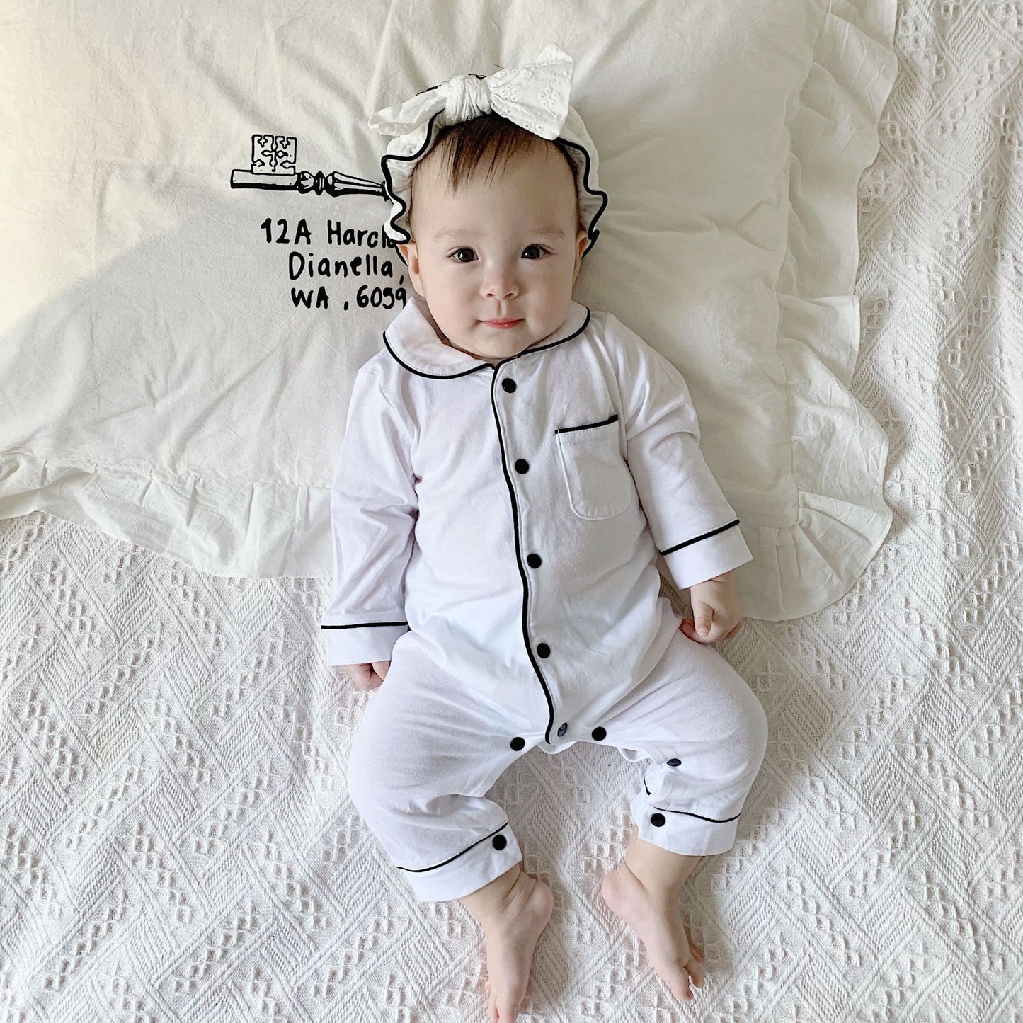 A baby in white pajamas with black trim and a matching headband lies on a patterned blanket, looking up with a slight smile. The pillow has whimsical text and a drawing.