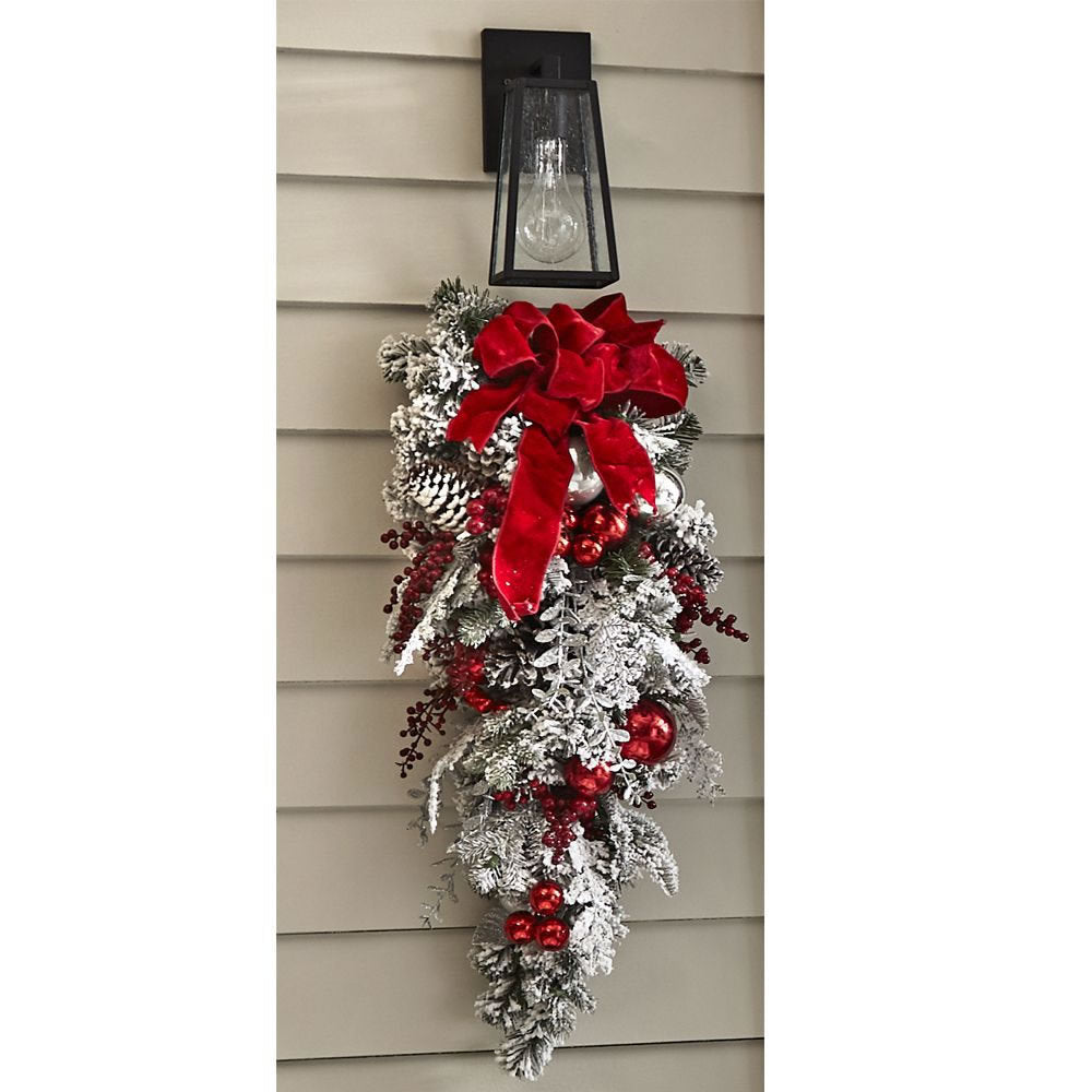 Festive holiday swag with frosted pinecones, red berries, and a large red ribbon, hanging under an outdoor lantern on a beige siding wall.