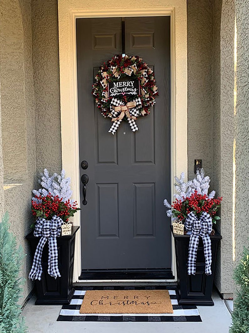 Festive front door with a "Merry Christmas" wreath, two planters with red berries and snowy branches, adorned with black and white checkered bows, and a "Merry Christmas" doormat.