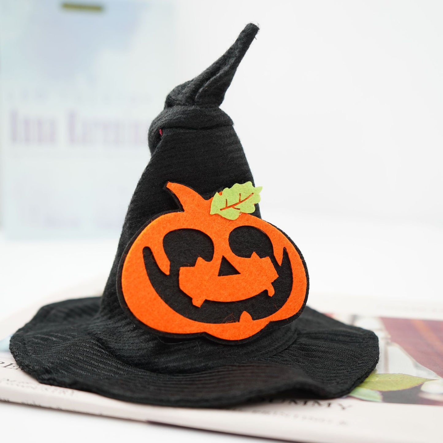 A black witch hat with a curled tip features a smiling orange felt pumpkin with a green leaf on a book background, creating a festive Halloween decoration.