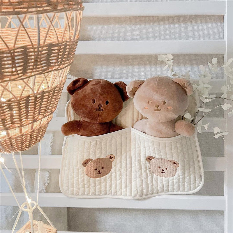 Two plush bears, one brown and one cream, sit in a white quilted hanging organizer with bear designs. A wicker lampshade and decorative branches complete the cozy nursery scene.