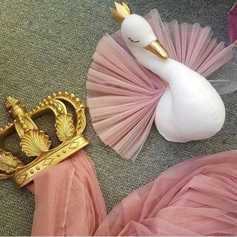 A plush white swan with a gold crown and pink tulle accents lies on a gray surface beside an ornate gold crown with pink fabric draped over one side.