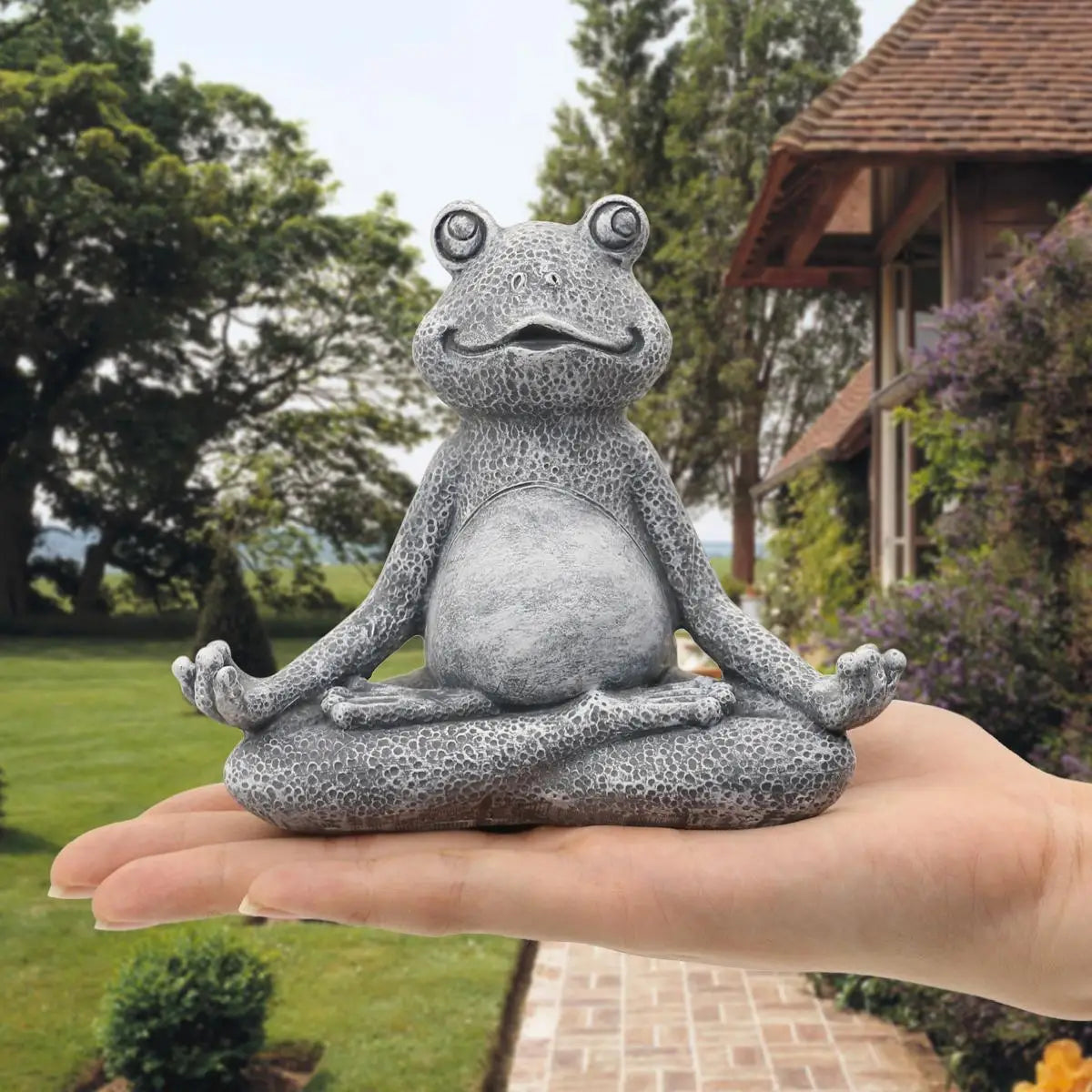 A small stone frog statue in a meditative pose is held in a hand, with a garden and house in the background. The statue has a textured surface and serene expression.