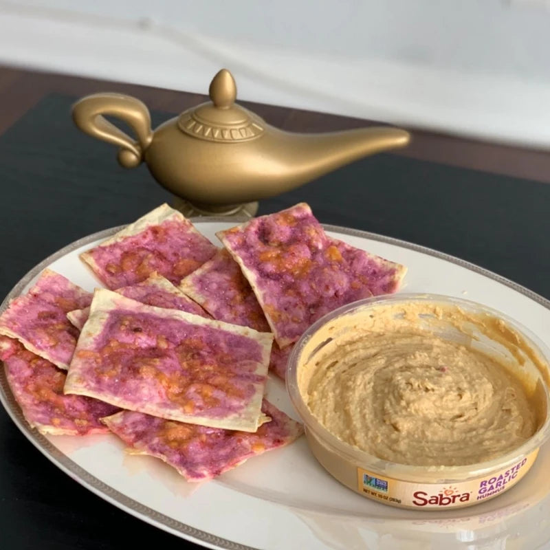 Plate with pink and orange flatbreads and a tub of roasted garlic hummus. A decorative, gold, genie-style lamp is placed in the background.