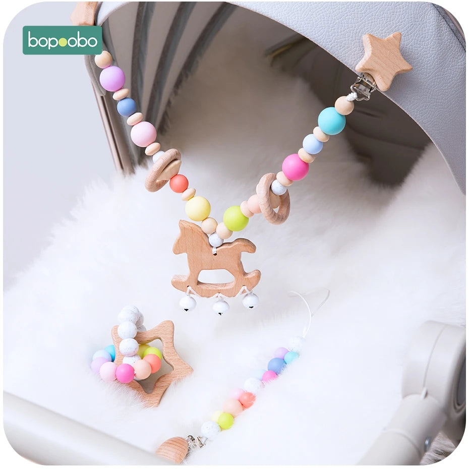 Colorful baby stroller chain with wooden beads, a horse-shaped pendant, and small bells. Includes a matching pacifier clip and a star-shaped rattle on a soft white background.