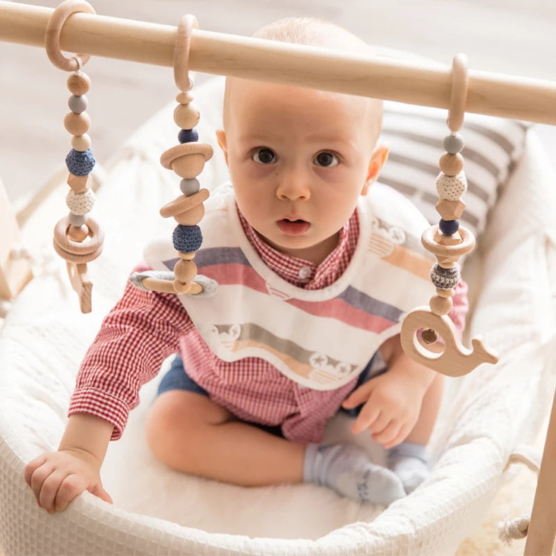 Baby sitting in a white bassinet, wearing a patterned bib and red checkered shirt, playing with a wooden hanging mobile with beads and a whale shape.