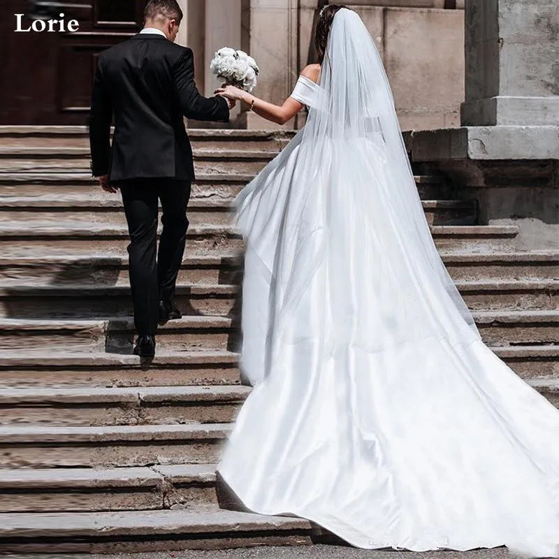 Bride in a flowing white gown and veil holds a bouquet, walking up stone steps with a groom in a black suit, hand in hand. Sunlight casts shadows on the steps.