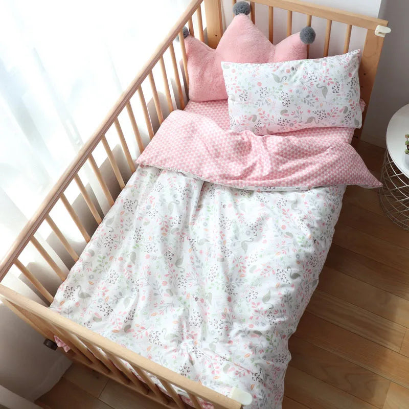 A wooden crib with a pink and white floral bedding set, featuring a pink crown-shaped cushion and pillow, sits on a wooden floor near a bright window.
