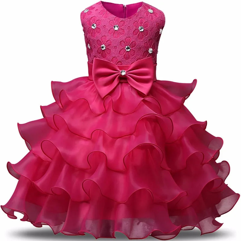 Pink sleeveless dress with a lace bodice adorned with small floral embellishments, a large bow at the waist, and a multi-layered ruffled skirt.