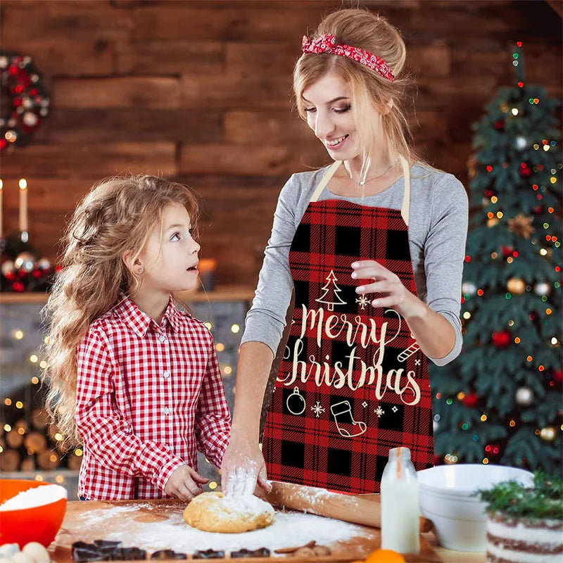 Woman in a "Merry Christmas" apron and young girl in plaid shirt baking together in a cozy, decorated kitchen with a Christmas tree and wreath in the background.
