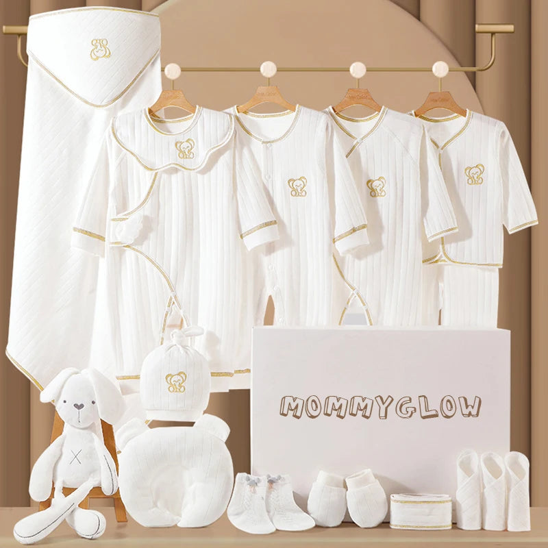A baby clothes set featuring white onesies, pajamas, and accessories with gold trim on hangers. Includes bathrobe, booties, a plush toy, and a box labeled "MommyGlow."