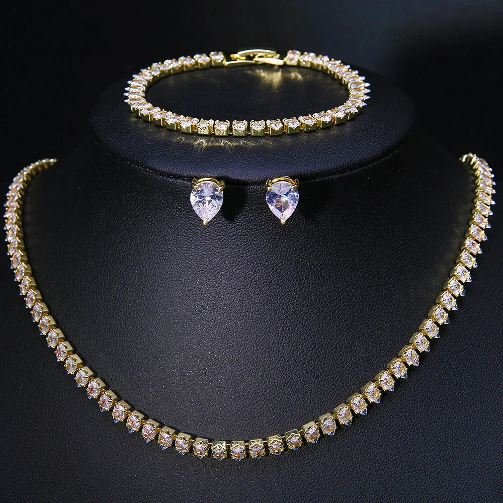 Gold jewelry set with sparkling white stones, including a necklace, bracelet, and teardrop earrings, displayed on a black stand against a dark background.