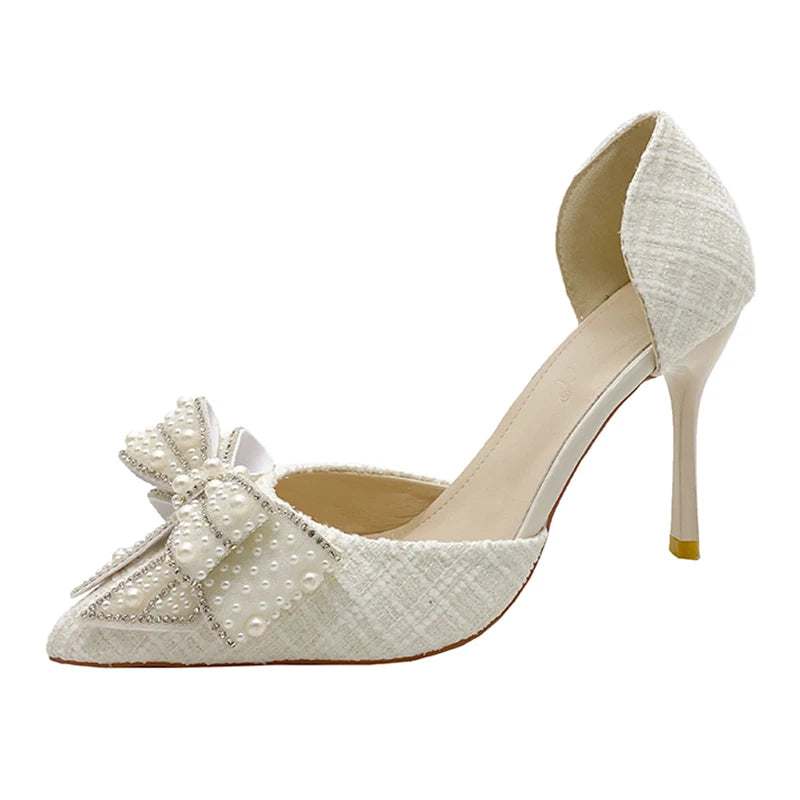 Elegant white high-heel shoe featuring a pointed toe, decorated with a large bow embellished with pearls and small crystals. The heel is slender and glossy.
