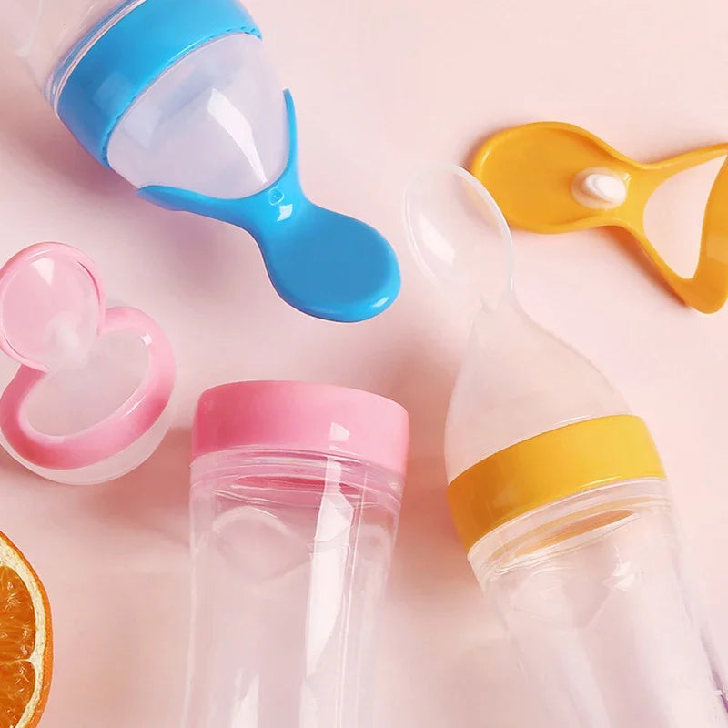 Colorful baby feeding bottles with silicone spoons and lids in pink, blue, and yellow on a light pink background. An orange slice is partially visible at the corner.