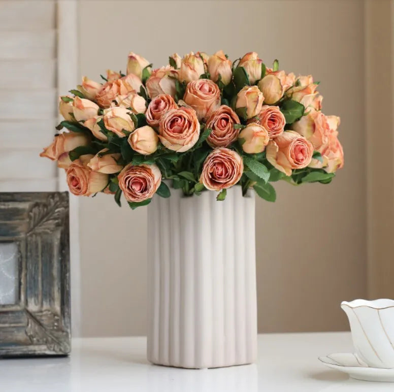A bouquet of peach roses in a tall, ribbed white vase on a table, next to a decorative frame and a white cup with a saucer.