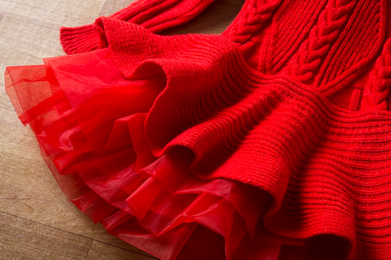 Close-up of a vibrant red sweater dress with intricate knit patterns and a flared, layered mesh skirt, laid flat on a wooden surface.