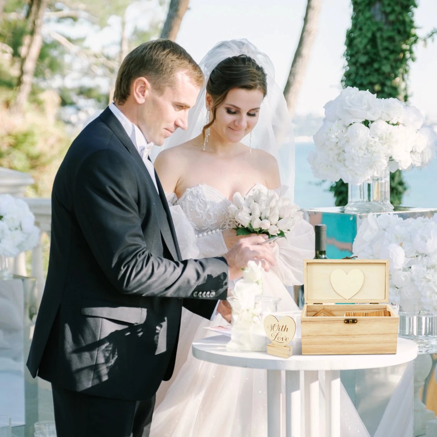 A bride and groom stand at a small table during their outdoor wedding, exchanging vows. The bride holds a bouquet of white flowers, and a wooden box with a heart design is open.