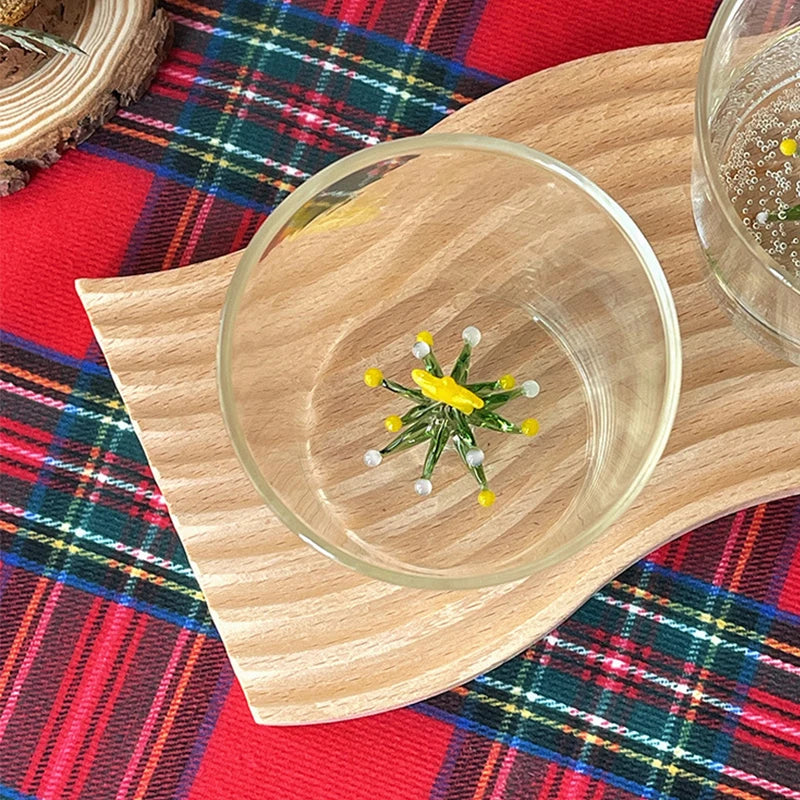 Glass cup with a floral stirrer on a wooden coaster, placed on a red tartan-patterned cloth. Another cup with bubbles is slightly visible on the right.