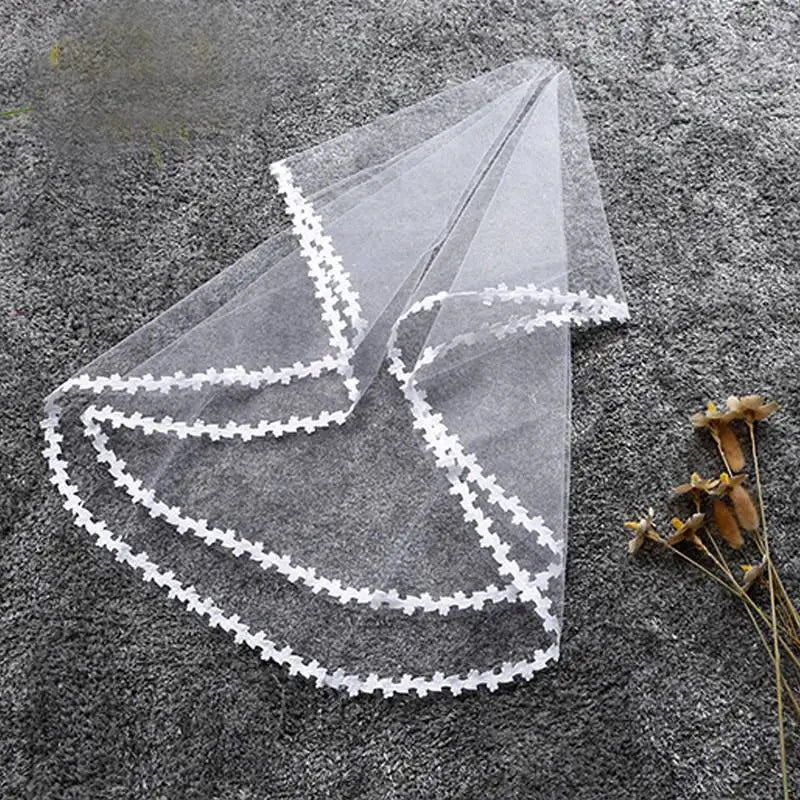 A translucent white veil with intricate border lace lays on a textured gray surface near dried flowers, creating a delicate and elegant composition.