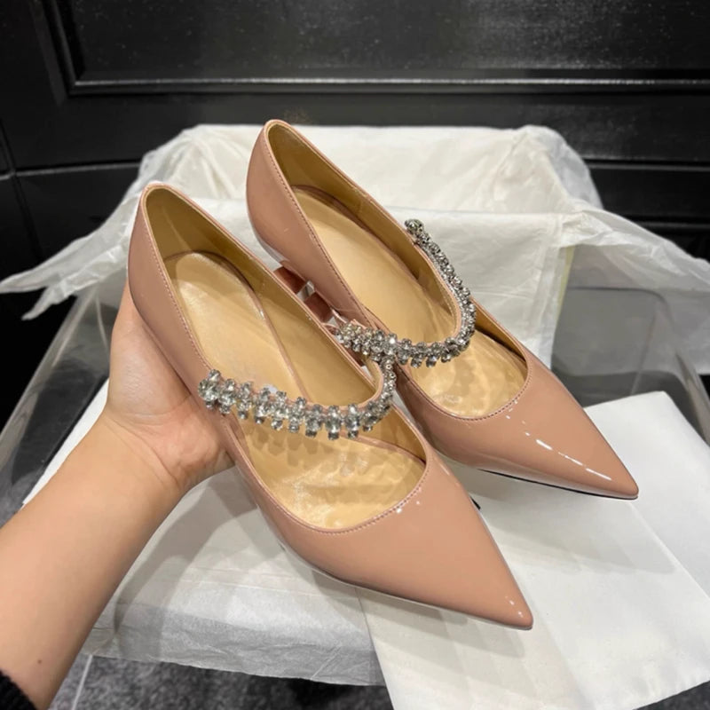 Beige pointed-toe heels with shiny rhinestone straps are displayed on a white box. A hand holds one shoe, revealing its sleek design and elegant embellishments.