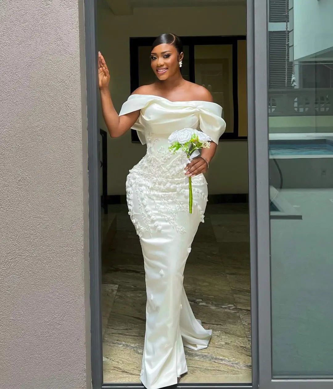 Woman in elegant off-shoulder white gown, smiling while holding a single white rose. She stands by a doorway, with sleek hair and pearl earrings enhancing her look.