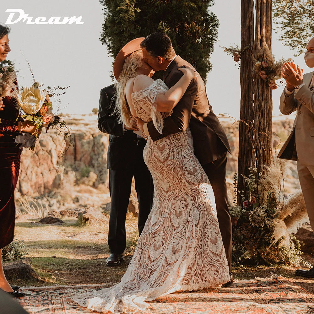 A couple shares a kiss during an outdoor wedding ceremony. The bride wears a lace gown and hat, while the groom is in a suit. Guests watch and applaud in a rustic, natural setting.