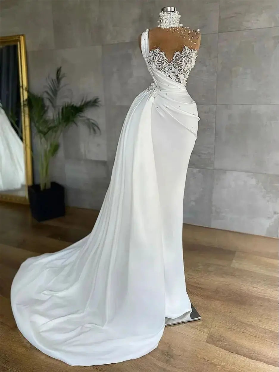 Elegant white gown on a mannequin with intricate lace bodice, pearl embellishments, and sweeping train. Set against a modern backdrop with a floor mirror and plant.