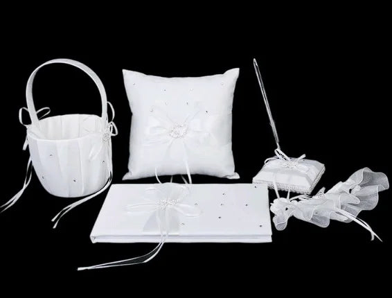 A white wedding set includes a flower basket, ring pillow, guest book with pen, garter, and two small pillows, embellished with bows and crystals, all against a black background.
