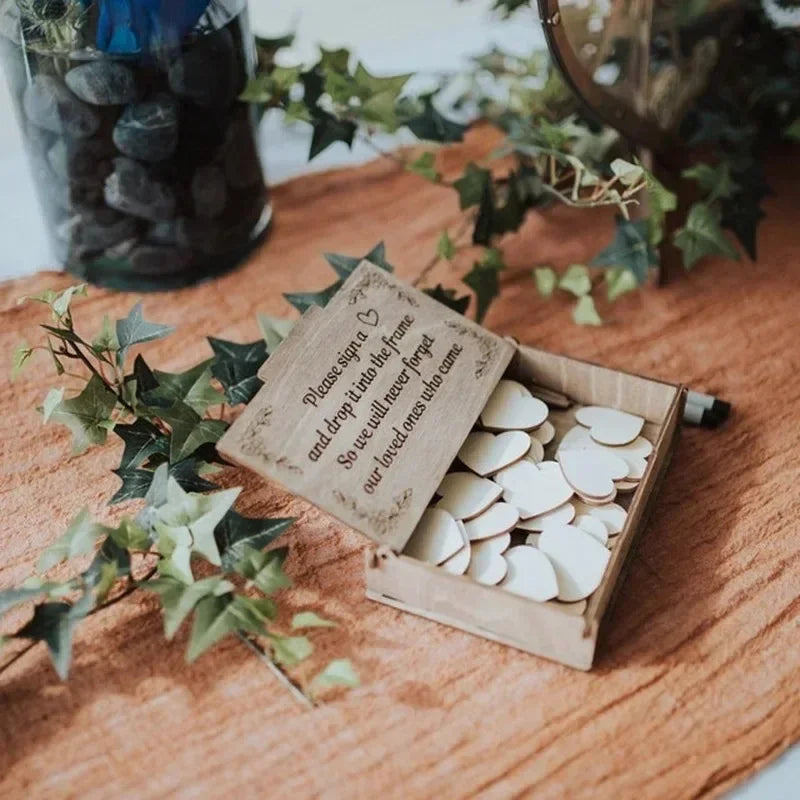 Wooden box with heart-shaped tokens and a small sign asking guests to sign for a keepsake. Surrounded by ivy decor on a textured brown surface.