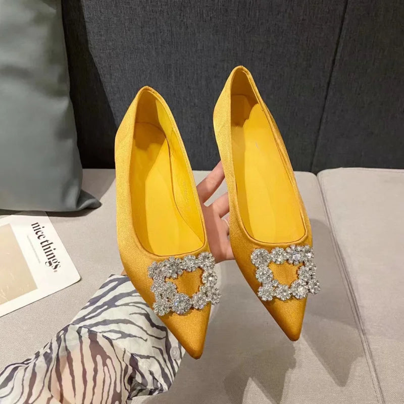 Yellow pointed-toe flats with sparkling crystal embellishments on the toes, held by a hand against a neutral background.