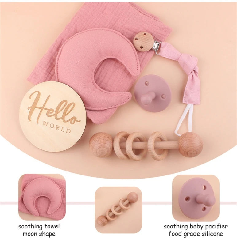 Baby gift set includes a wooden "Hello World" sign, pink moon-shaped towel, silicone pacifier, wooden rattle, and a pacifier clip. Items in muted pink and natural wood tones.