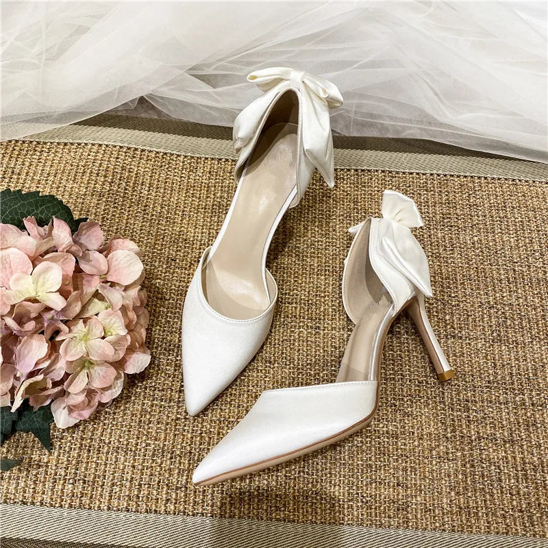 Elegant white high heels with bows, resting on a textured brown mat. A bouquet of soft pink hydrangeas is placed nearby, with sheer white fabric in the background.