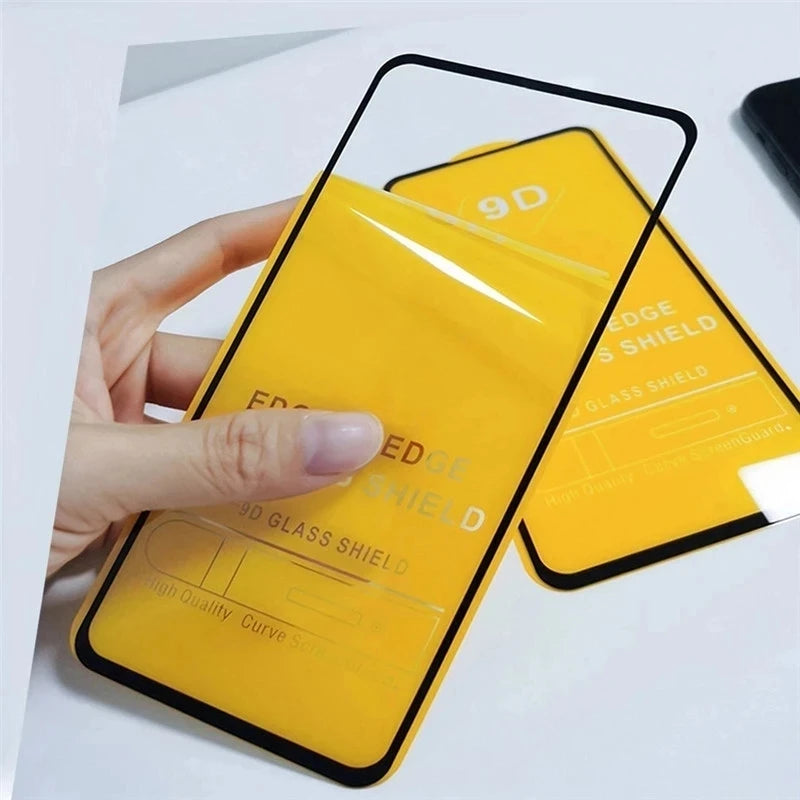 A hand holds a transparent, curved-edge 9D glass screen protector with a black border, against a yellow backing. Another protector lies beneath on a white surface.