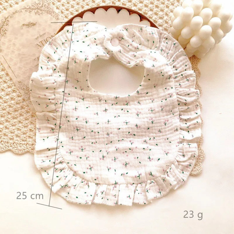 White, ruffled baby bib with small floral pattern, dimensions 25 cm and weight 23 g. The bib is laid on a textured mat with a decorative white candle nearby.