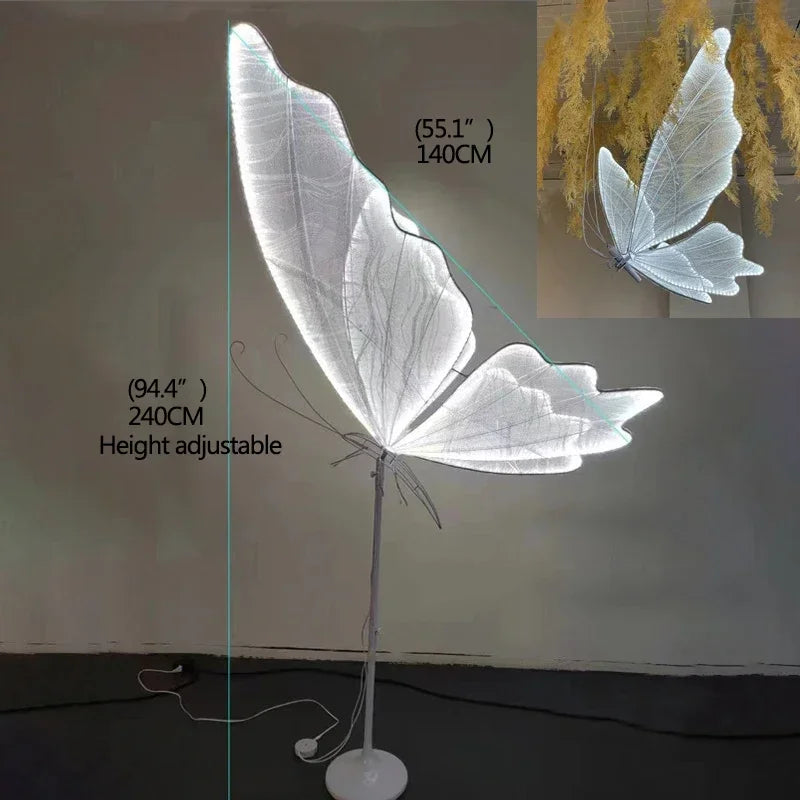 Butterfly-shaped floor lamp with glowing white wings, height adjustable from 55.1" to 94.4" (140-240cm). Inset shows lamp hanging amid dry foliage.