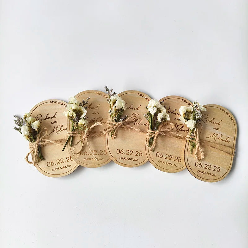 Wooden wedding save-the-date magnets, decorated with small white flowers and twine, announce a June 22, 2025, event in Oakland, CA. Five are arranged in a row.