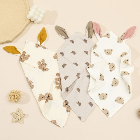 Three baby cloths with playful designs: one with teddy bears and mustard ears, one with rainbows and grey ears, and one with bear faces and pink ears, laid on a beige surface.